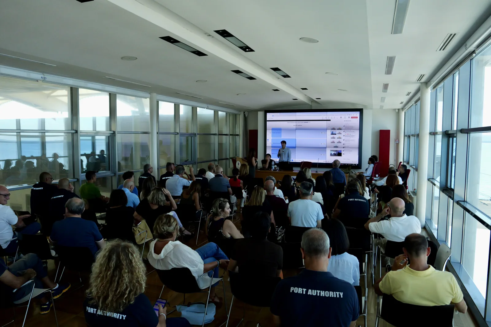 Plenaria affollata davanti al maxischermo, in una sala con le vetrate sul mare